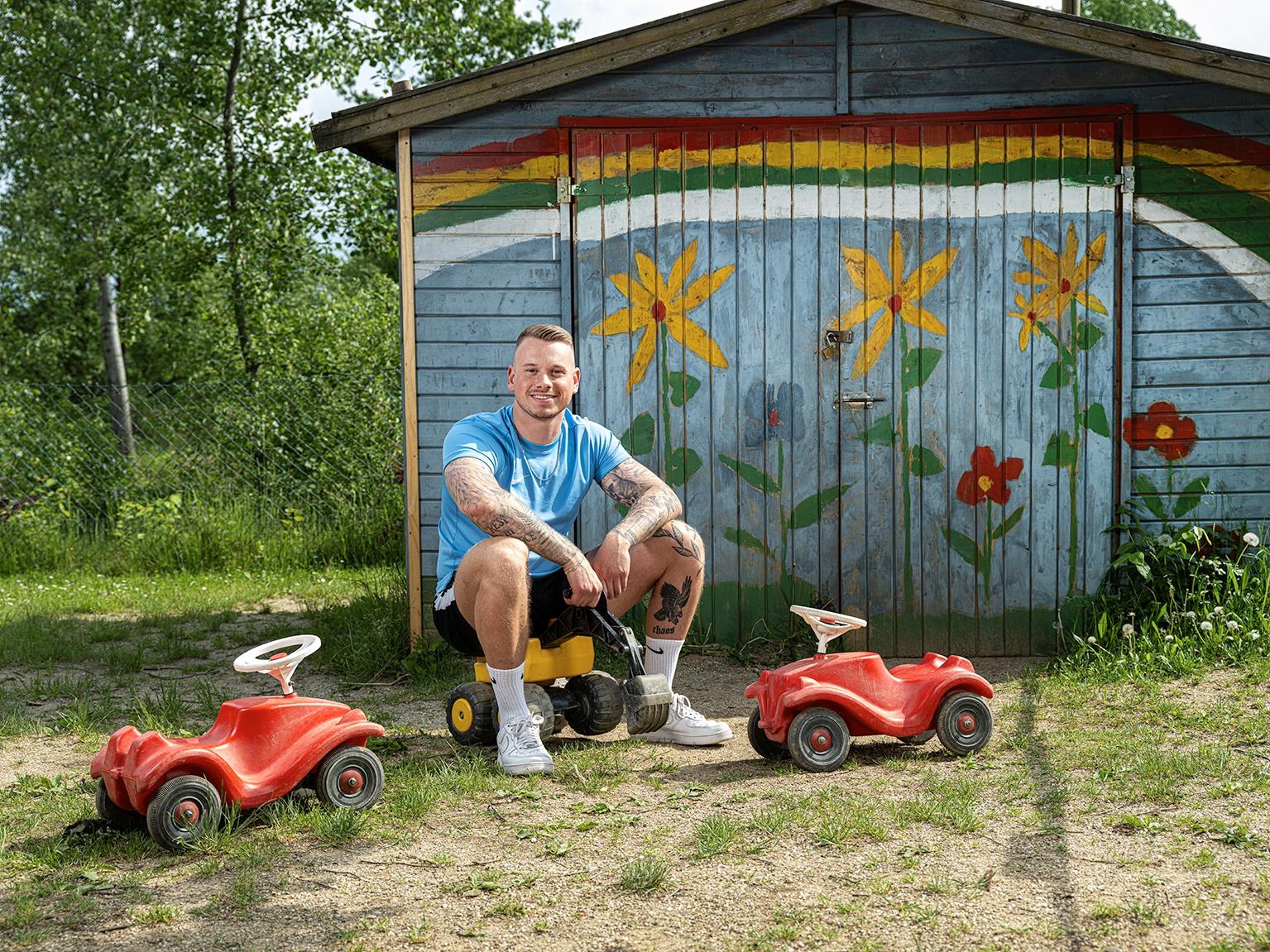 Junger Mann sitzt lachend vor einem bunt angemalten Schuppen zwischen Bobby Cars da.